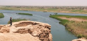 Person standing on a dry rocky soil, overlooking a river with city in the background