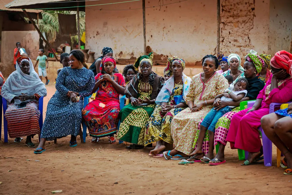 Group of women sitting on chairs in a row outside, wearing colourful clothes